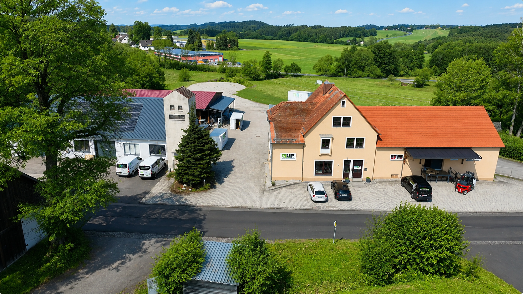 Luftaufnahme eines Firmengebäudes mit Lager- und Betriebsflächen in ländlicher Umgebung. Das Gelände ist von grünen Wiesen, Bäumen und Feldern umgeben und zeigt den Standort an einem sonnigen Sommertag aus der Vogelperspektive.